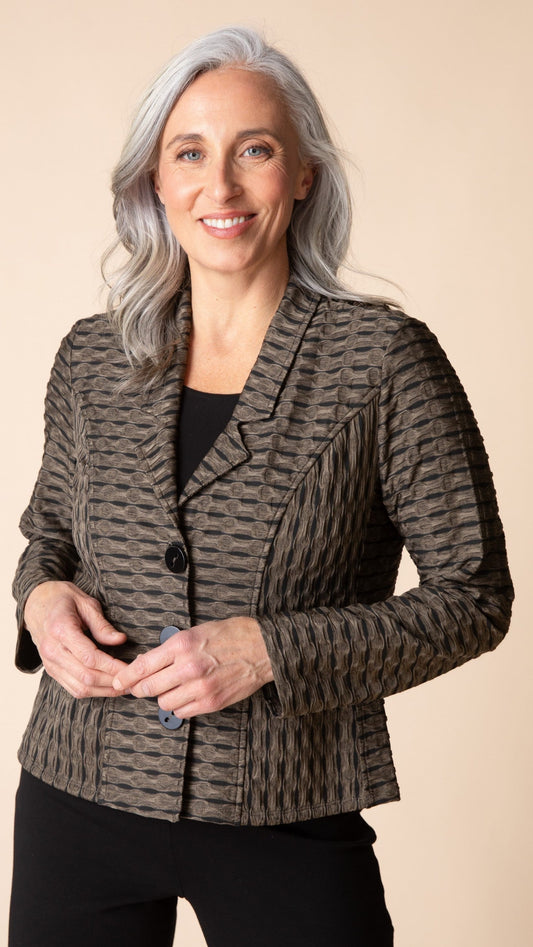 A smiling woman with gray hair wearing a textured, patterned brown and gray blazer over a black top, standing against a solid beige background.