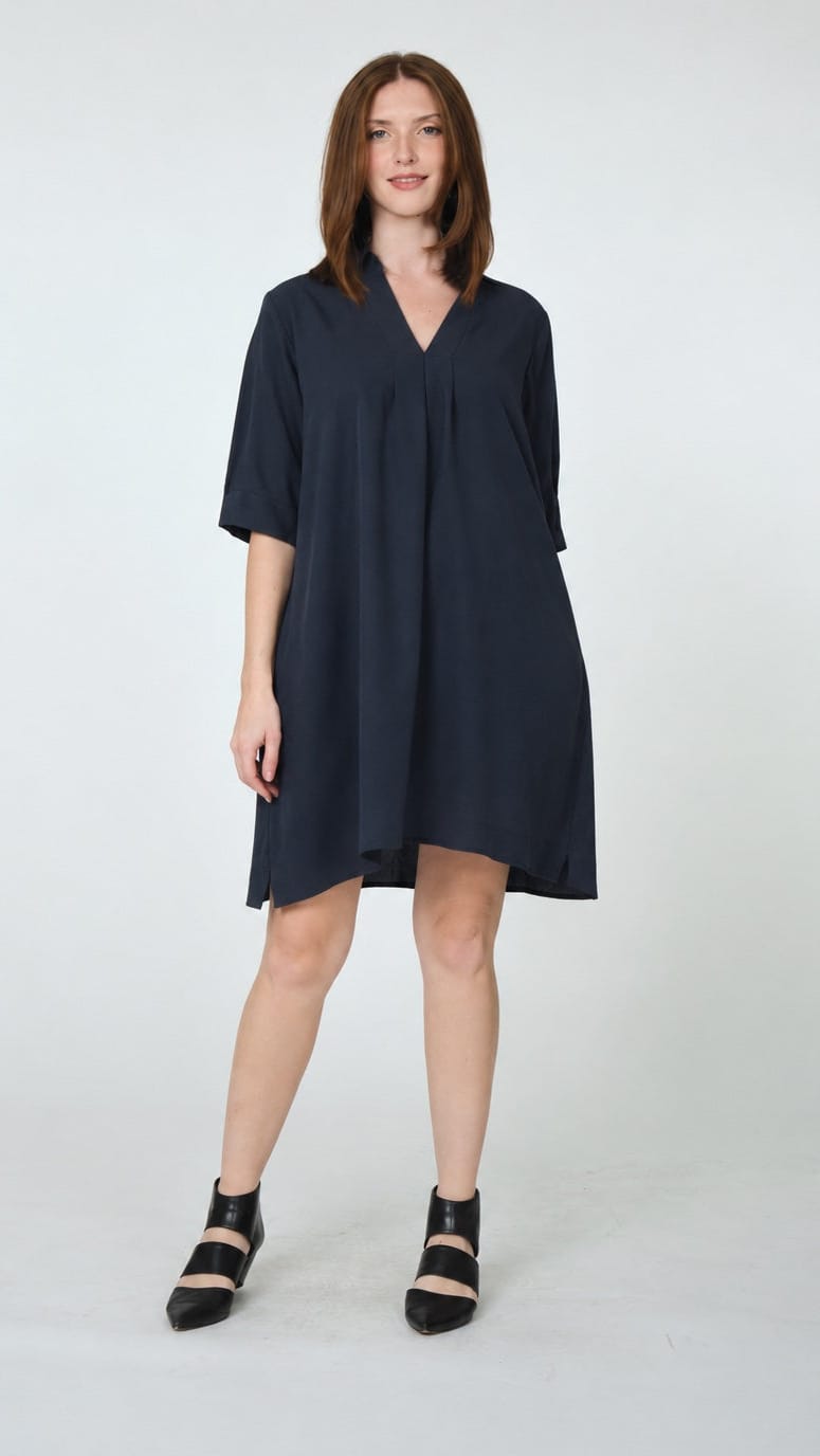 A woman modeling a loose-fitting navy blue short-sleeve V-neck dress with a gathered front, paired with black strappy heels, against a light gray studio background.