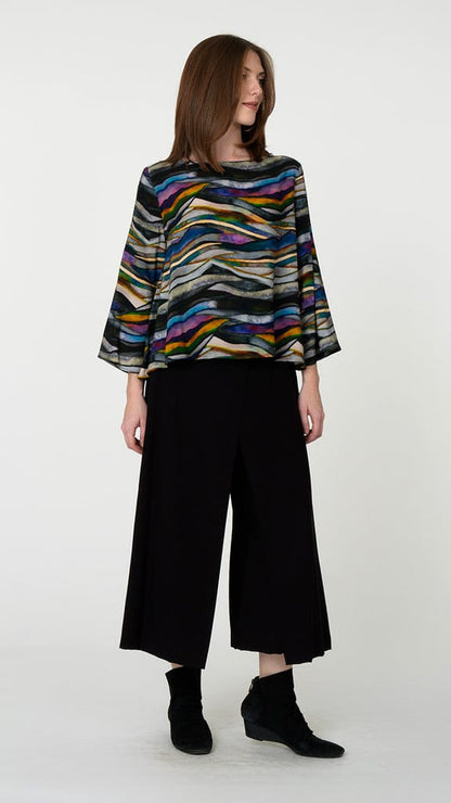 A woman modeling a colorful, wide-sleeved top with a wavy multicolor stripe pattern, paired with black wide-leg cropped pants and black ankle boots, against a plain light gray background.