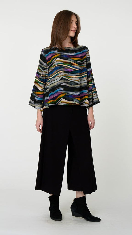 A woman modeling a colorful, wide-sleeved top with a wavy multicolor stripe pattern, paired with black wide-leg cropped pants and black ankle boots, against a plain light gray background.