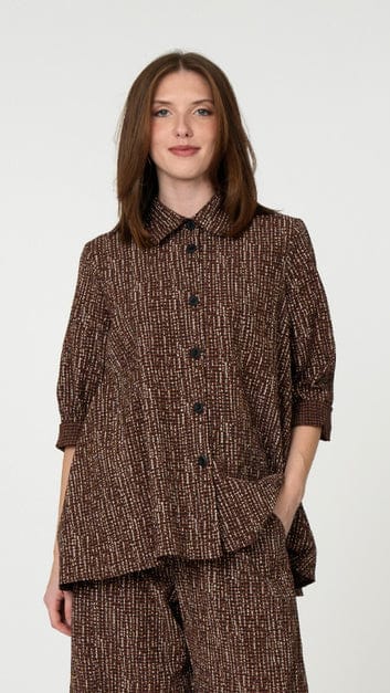 A woman wearing a brown and beige textured tweed button-up shirt and matching trousers, photographed against a plain light gray studio background.