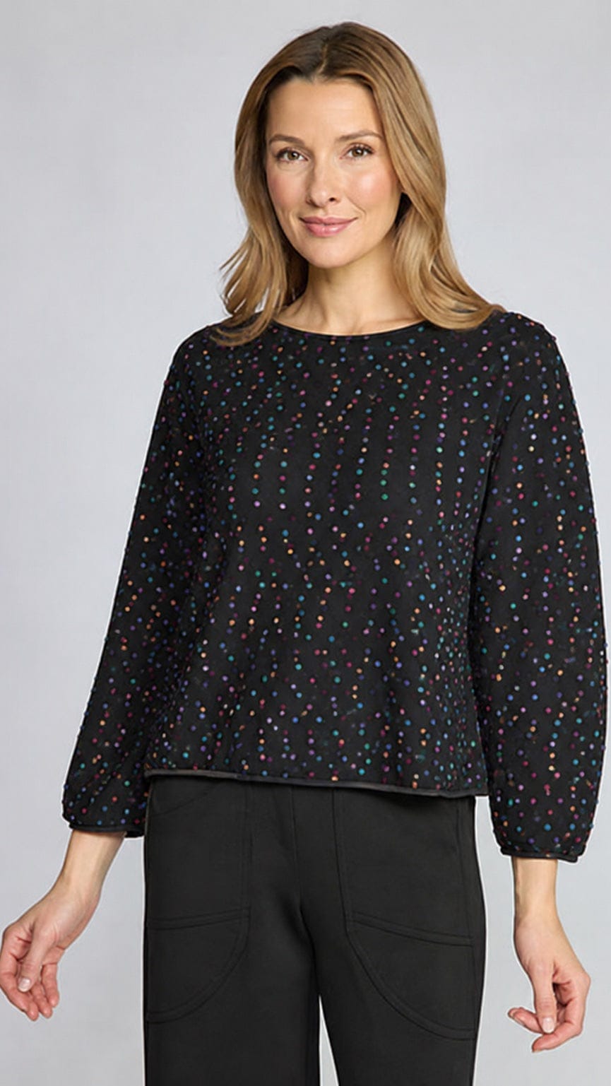 A woman modeling a black long-sleeve top with a small multicolor polka dot pattern, paired with black pants, against a solid light gray background.