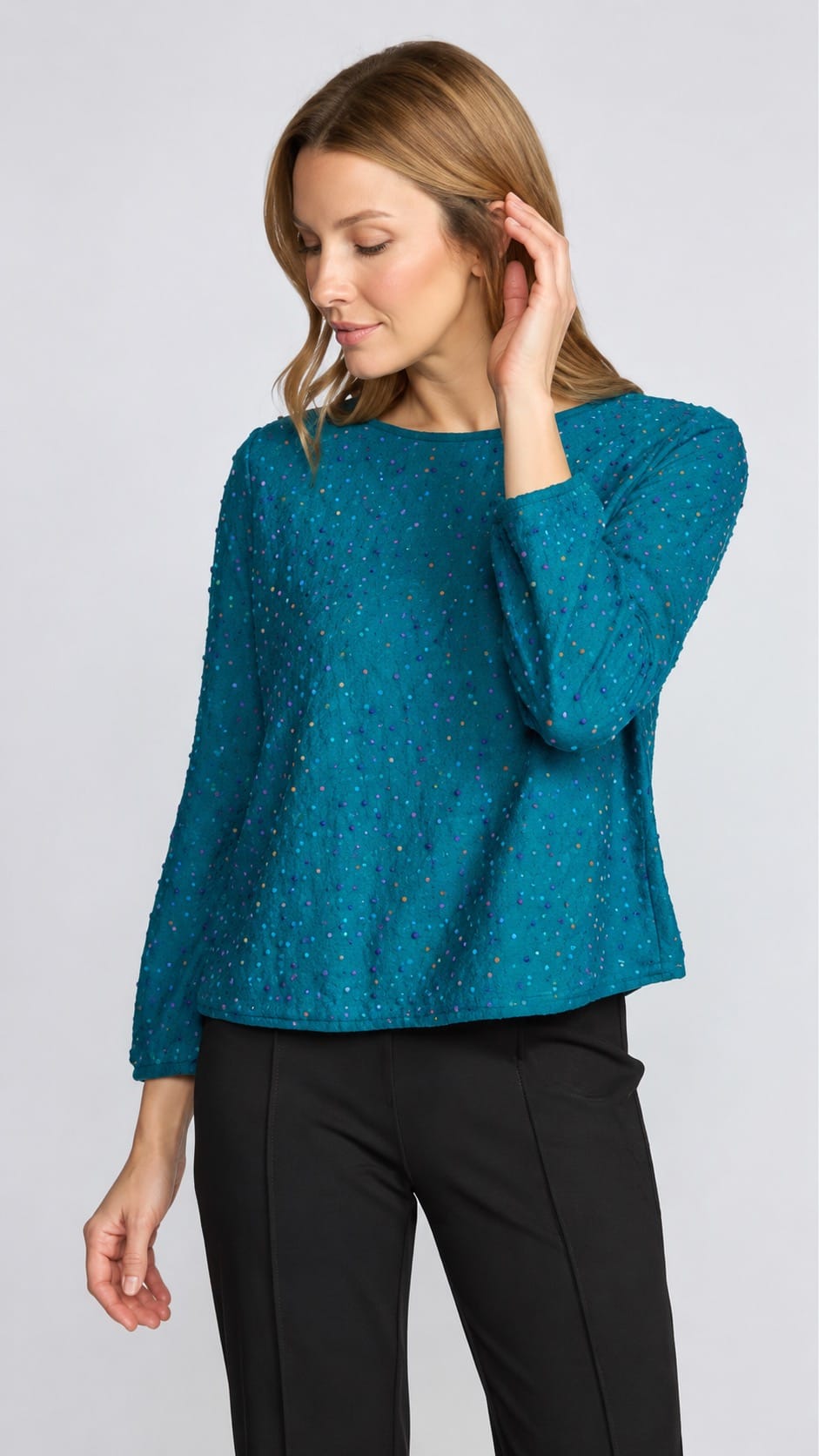 A woman wearing a teal blue textured knit sweater with three-quarter sleeves and black pants, standing against a light gray studio background.