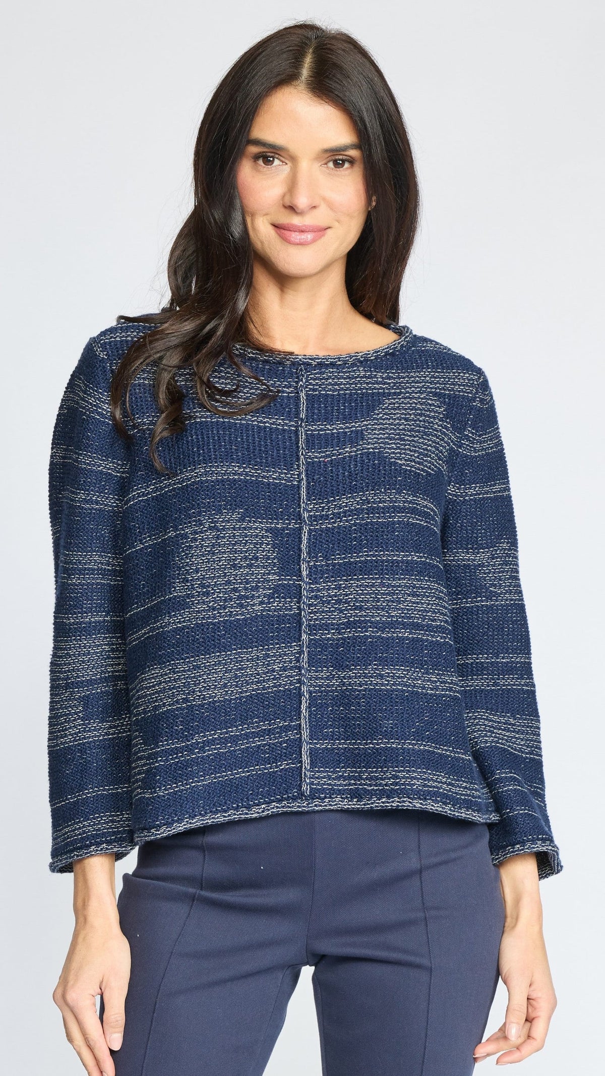 A woman modeling a textured knit sweater with a blue and gray marled pattern, paired with navy pants, against a light gray studio background.