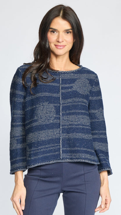 A woman modeling a textured knit sweater with a blue and gray marled pattern, paired with navy pants, against a light gray studio background.