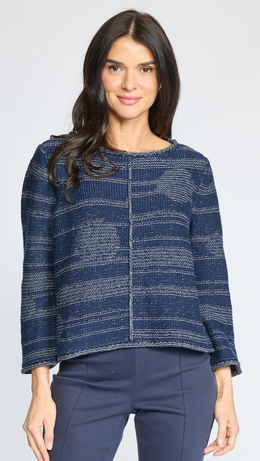 A woman modeling a textured knit sweater with a blue and gray marled pattern, paired with navy pants, against a light gray studio background.