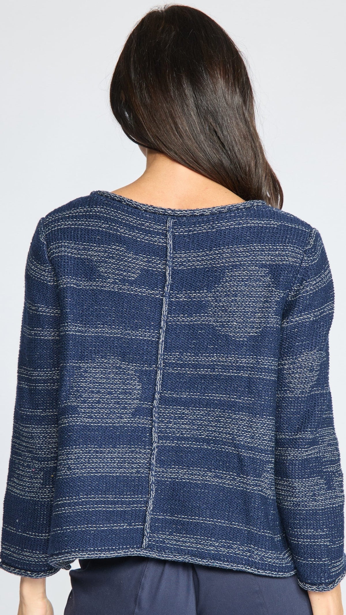 A woman viewed from the back wearing a textured navy blue knit sweater with horizontal stripe patterns and a central seam, paired with dark blue pants against a light gray background.