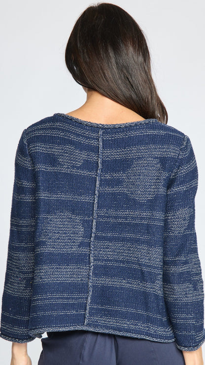A woman viewed from the back wearing a textured navy blue knit sweater with horizontal stripe patterns and a central seam, paired with dark blue pants against a light gray background.