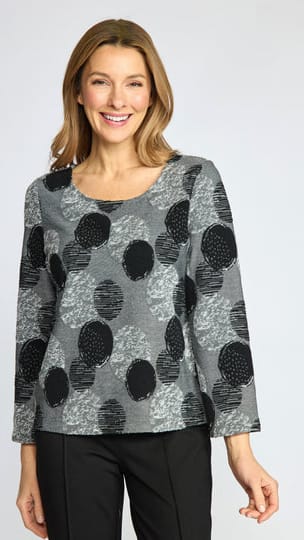 A woman wearing a gray long-sleeve top with a black and gray abstract polka dot pattern, paired with black pants, against a light gray studio background.