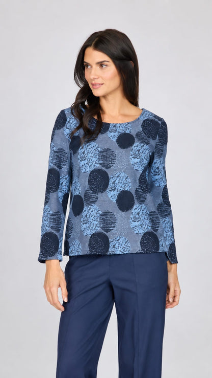 A woman modeling a long-sleeve blouse with a blue and navy abstract circular pattern, paired with navy trousers, against a light gray studio background.