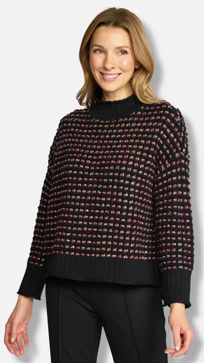 A woman wearing a black knit sweater with a textured multicolor pattern of small pink, red, and green accents, paired with black pants, against a light gray studio background.