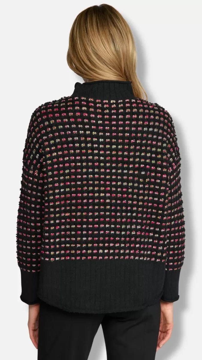 A woman viewed from the back wearing a black knit sweater with a textured multicolor grid pattern of pink, red, and green threads, paired with black pants, against a light gray background.