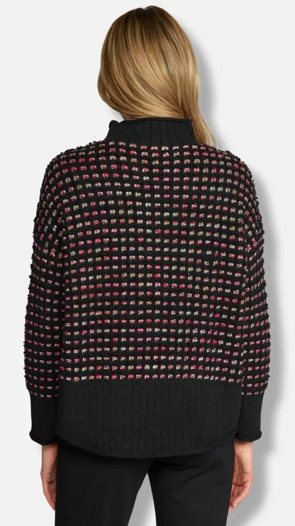 A woman viewed from the back wearing a black knit sweater with a textured multicolor grid pattern of pink, red, and green threads, paired with black pants, against a light gray background.