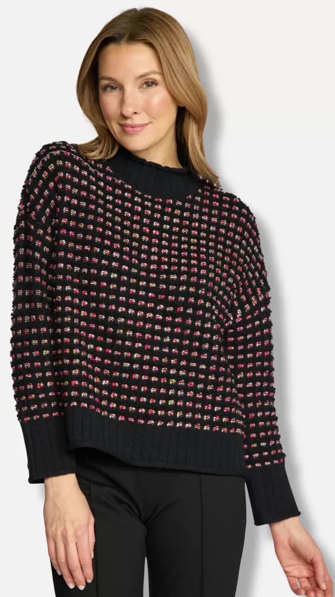 A woman wearing a black knit sweater with a textured multicolor tweed pattern of pink, red, and beige threads, paired with black pants, against a light gray studio background.