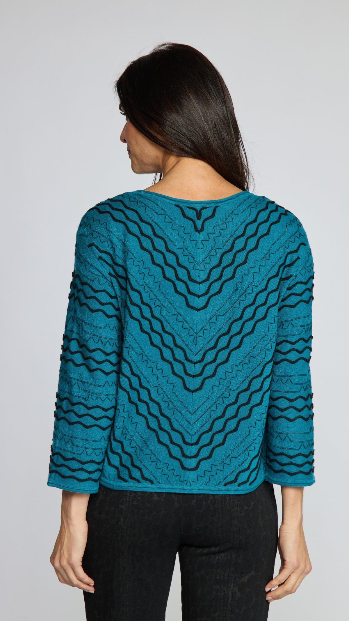 A woman modeling the back of a teal long-sleeve knit top with a black wavy geometric pattern, paired with black pinstriped trousers, against a solid gray studio background.