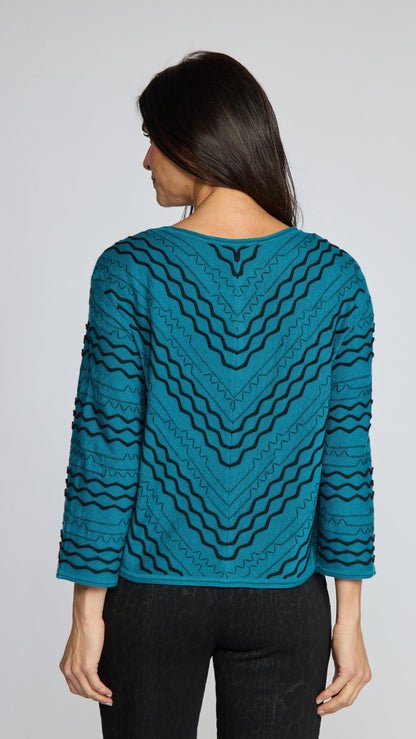 A woman modeling the back of a teal long-sleeve knit top with a black wavy geometric pattern, paired with black pinstriped trousers, against a solid gray studio background.