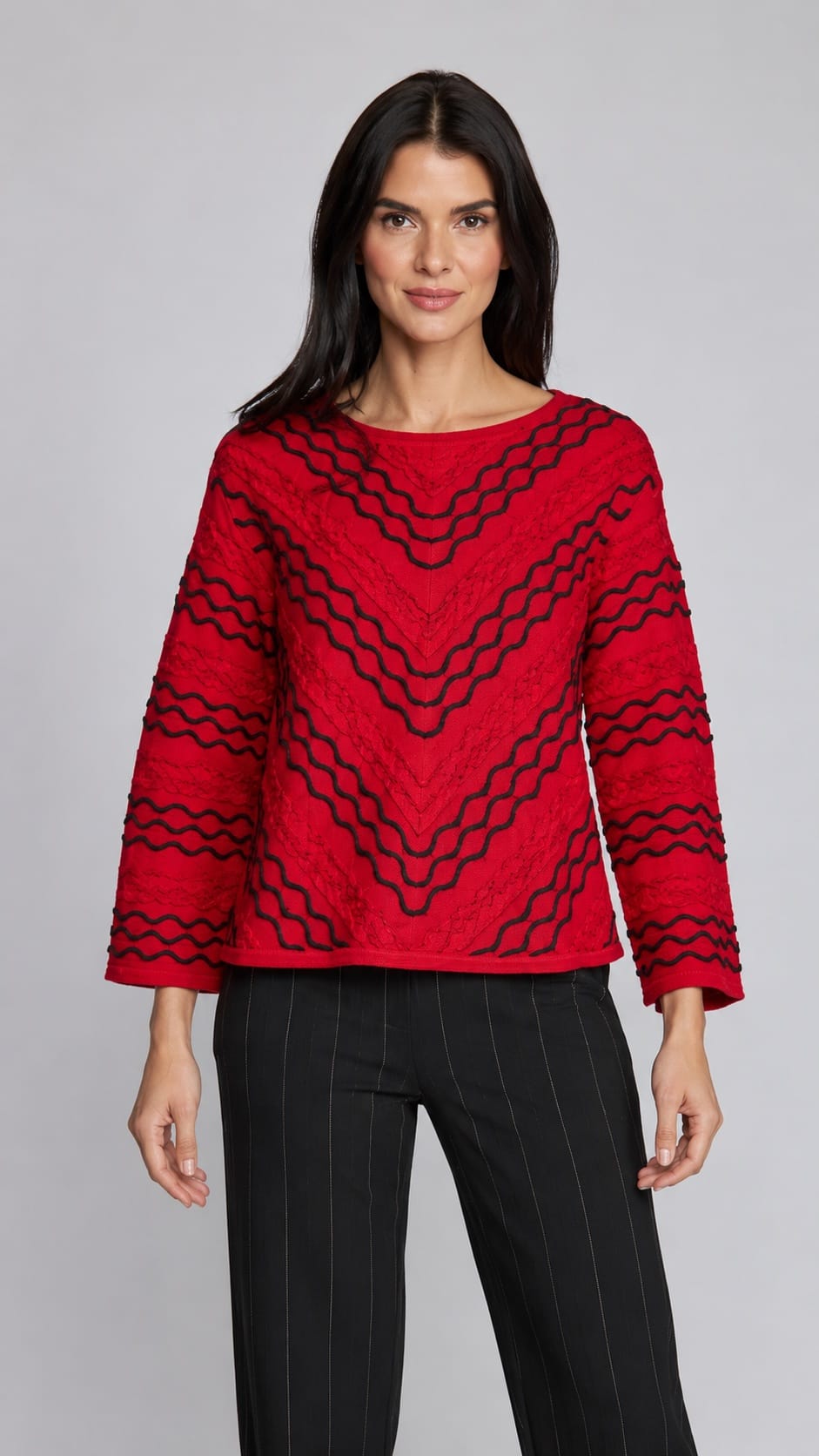 A woman modeling a red long-sleeve knit top with a black wavy geometric pattern, paired with black pinstriped trousers, against a solid gray studio background.