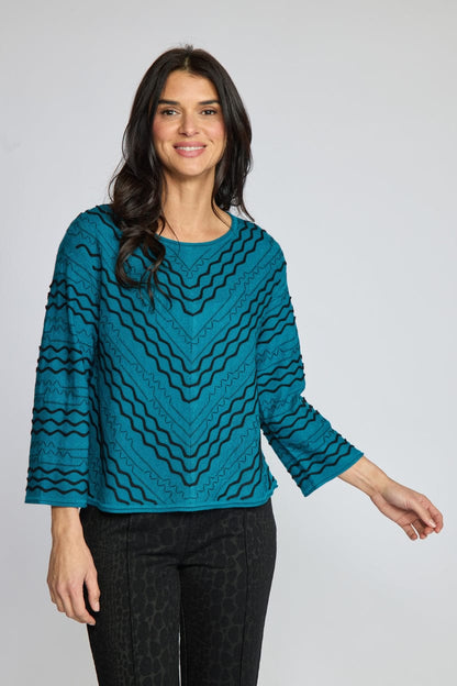 A woman modeling a teal long-sleeve knit top with a black wavy geometric pattern, paired with black pinstriped trousers, against a solid gray studio background.