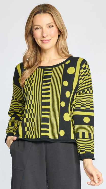 A woman modeling a long-sleeve knit top with a bold black and lime green geometric pattern, paired with dark gray pants, against a light gray studio background.