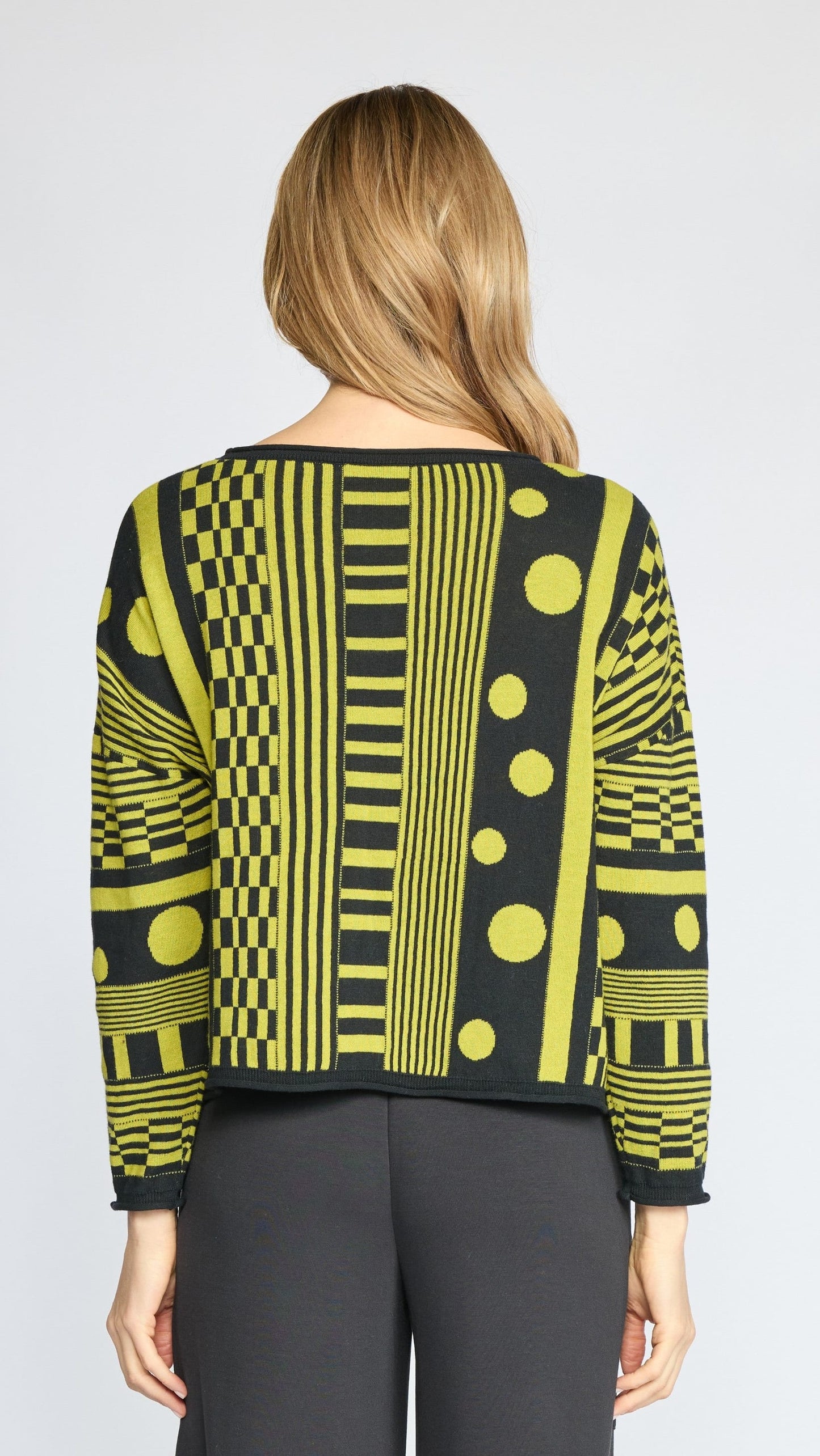 A woman viewed from the back wearing a long-sleeve knit top with a bold yellow and black geometric pattern, paired with dark gray pants, against a light gray studio background.