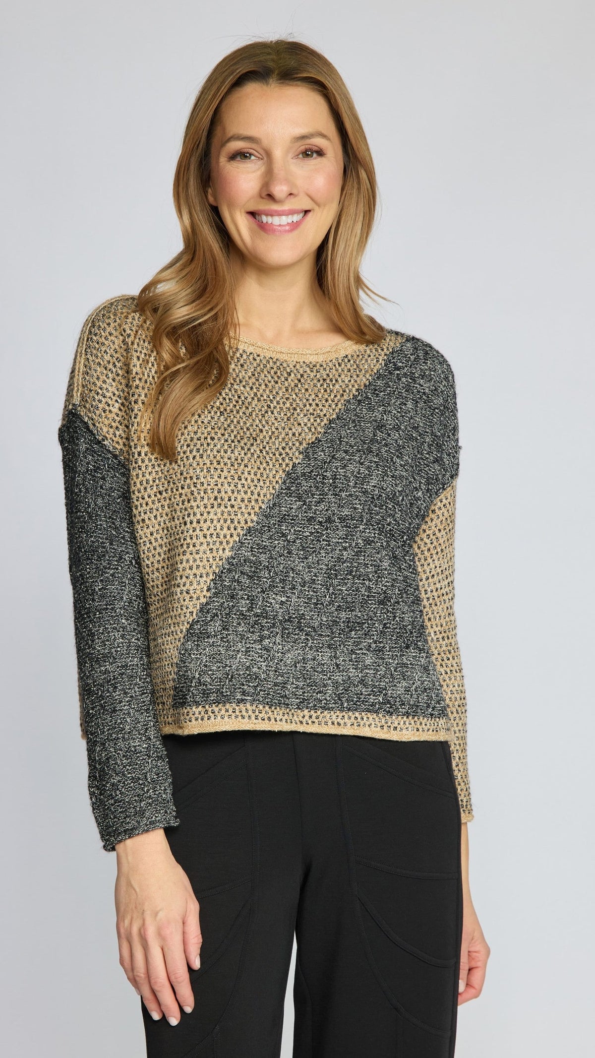 A woman modeling a color-block knit sweater with a beige and dark gray pattern, paired with black pants, against a light gray studio background.