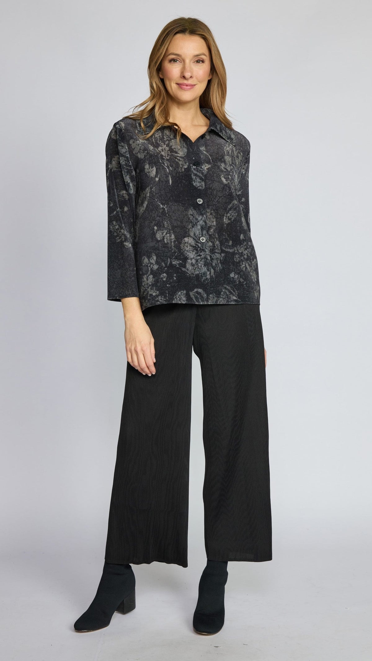 A woman modeling a dark gray and black floral patterned button-up shirt with three-quarter sleeves, paired with black wide-leg trousers and black ankle boots, against a light gray studio background.