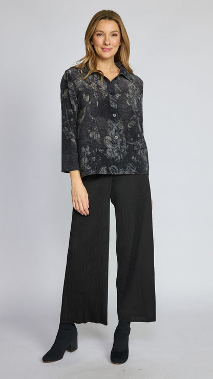 A woman modeling a dark gray and black floral patterned button-up shirt with three-quarter sleeves, paired with black wide-leg trousers and black ankle boots, against a light gray studio background.