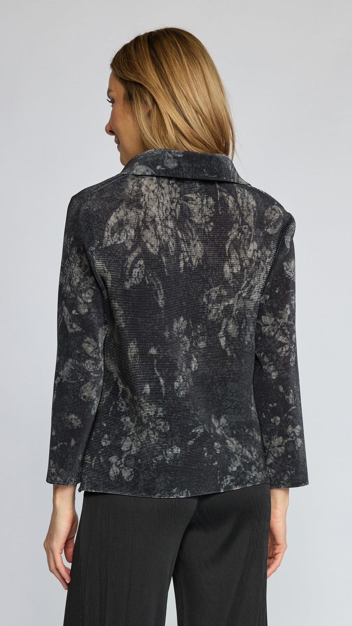 A woman shown from the back wearing a dark charcoal gray textured blazer with a lighter gray abstract floral pattern, paired with black trousers, against a light gray studio background.