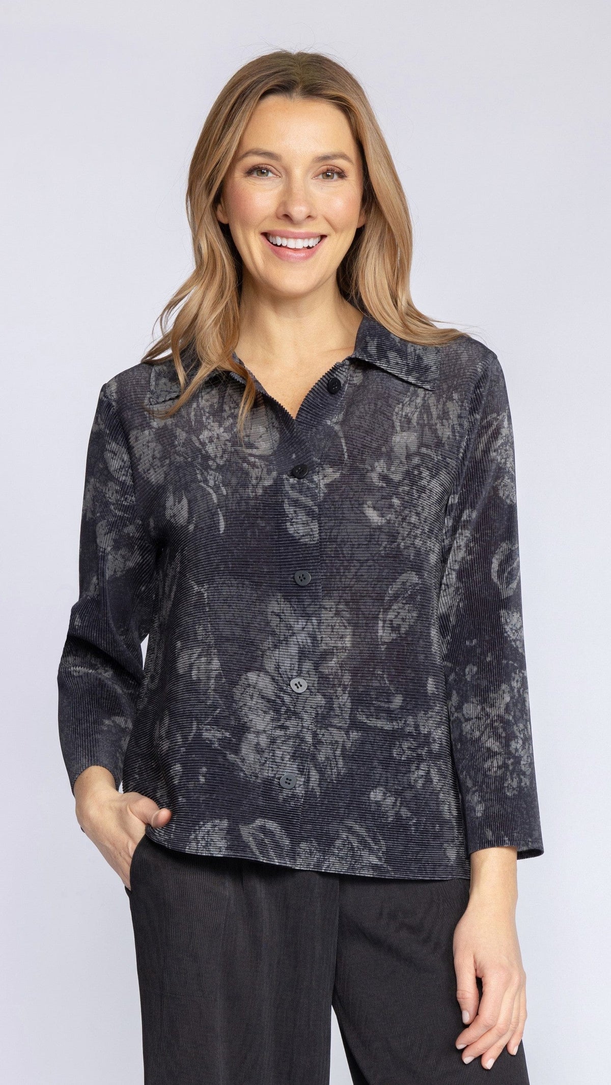 A woman modeling a long-sleeve button-up shirt with a dark gray and black abstract floral pattern, paired with black pants, against a light gray studio background.