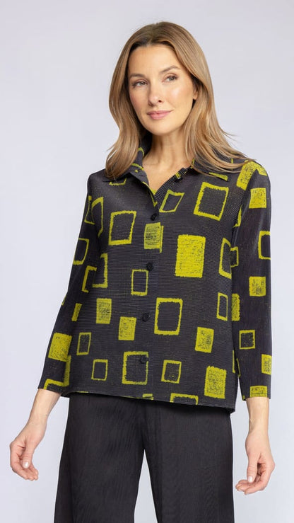 A woman modeling a black long-sleeve button-up shirt with a bright yellow-green abstract geometric pattern of rectangles, paired with black trousers, against a light gray studio background.