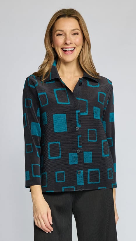 A smiling woman wearing a black button-up shirt with a teal geometric square pattern, paired with dark pants, against a light gray studio background.