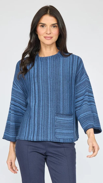 A woman modeling a blue striped knit top with three-quarter sleeves and a front patch pocket, paired with navy pants, against a light gray studio background.
