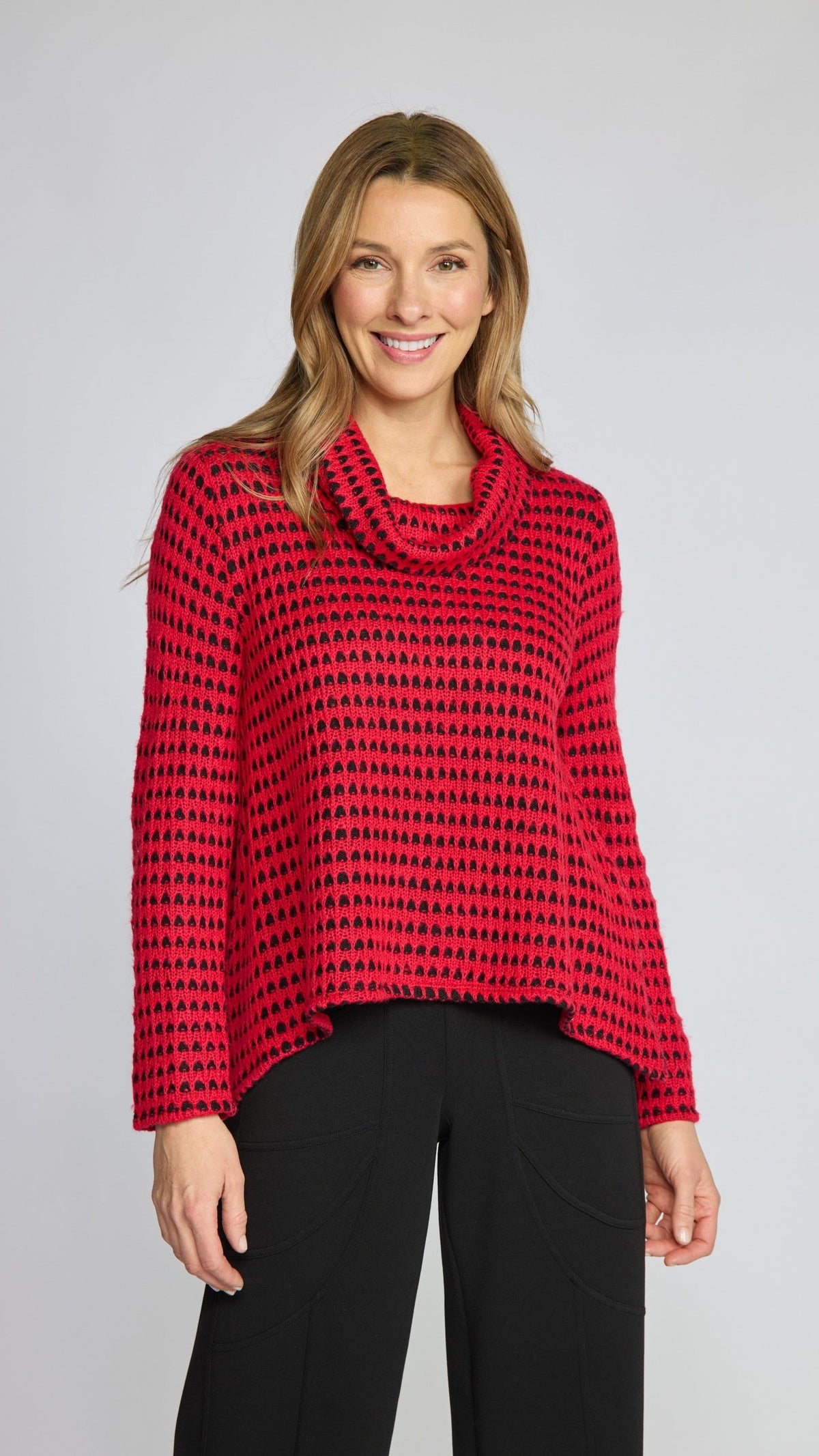 A woman modeling a red and black patterned knit cowl-neck sweater with black pants, photographed against a light gray studio background.