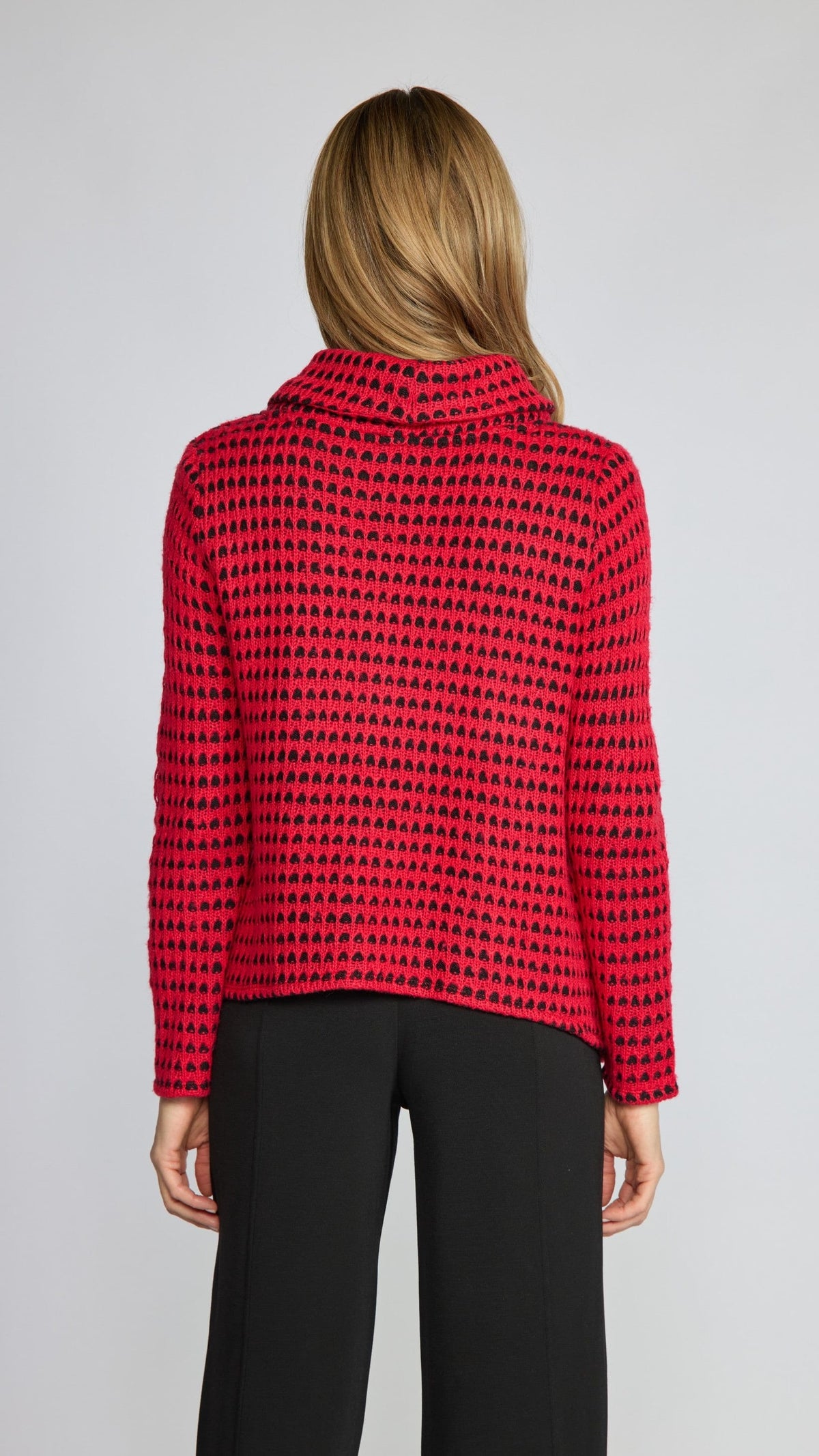 A woman viewed from the back wearing a red knit long-sleeve top with a black geometric pattern and black trousers, against a light gray studio background.