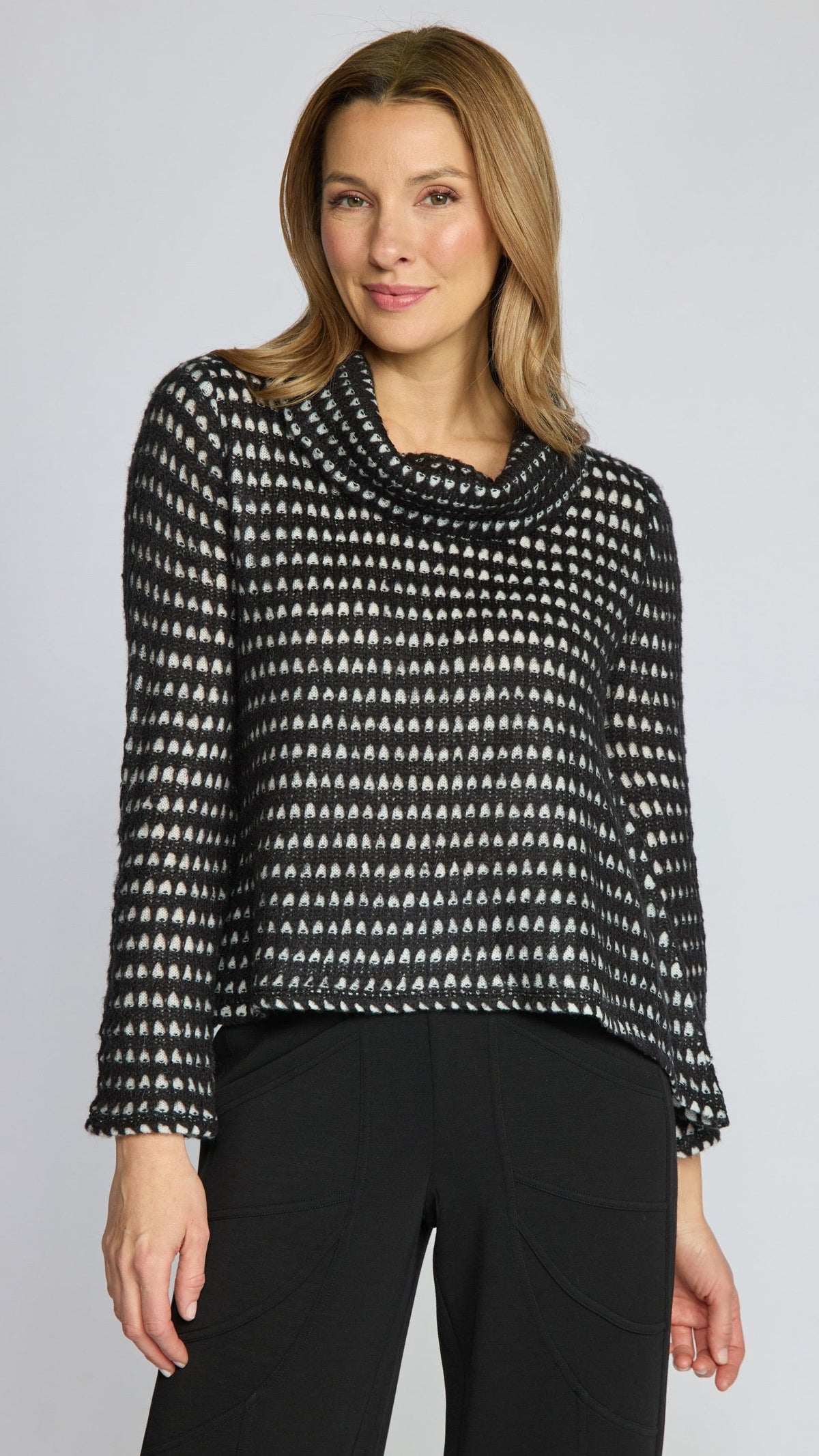 A woman modeling a black and white patterned knit cowl-neck top with long sleeves, paired with black pants, against a light gray studio background.