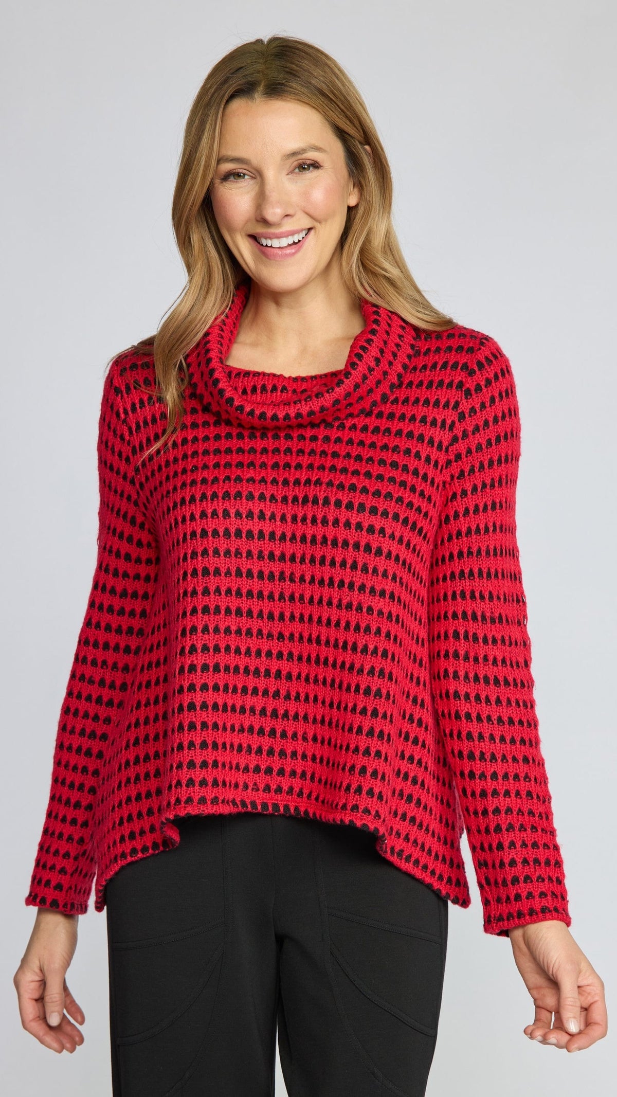A woman modeling a red and black patterned knit cowl-neck sweater with black pants, photographed against a light gray studio background.