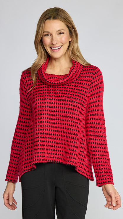 A woman modeling a red and black patterned knit cowl-neck sweater with black pants, photographed against a light gray studio background.