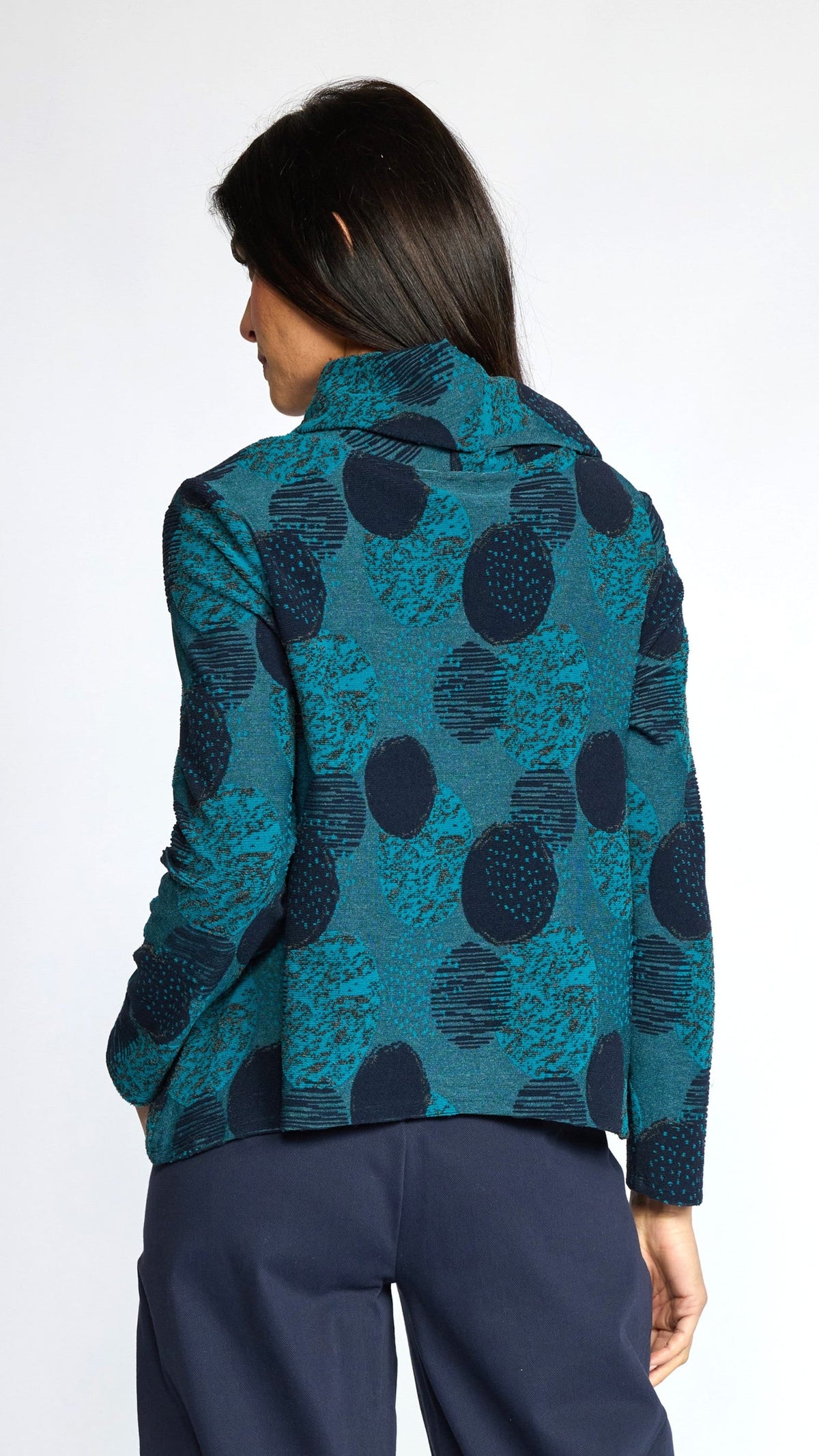 A woman shown from the back wearing a teal and navy patterned blazer with large abstract circular motifs, paired with navy trousers, against a plain white studio background.