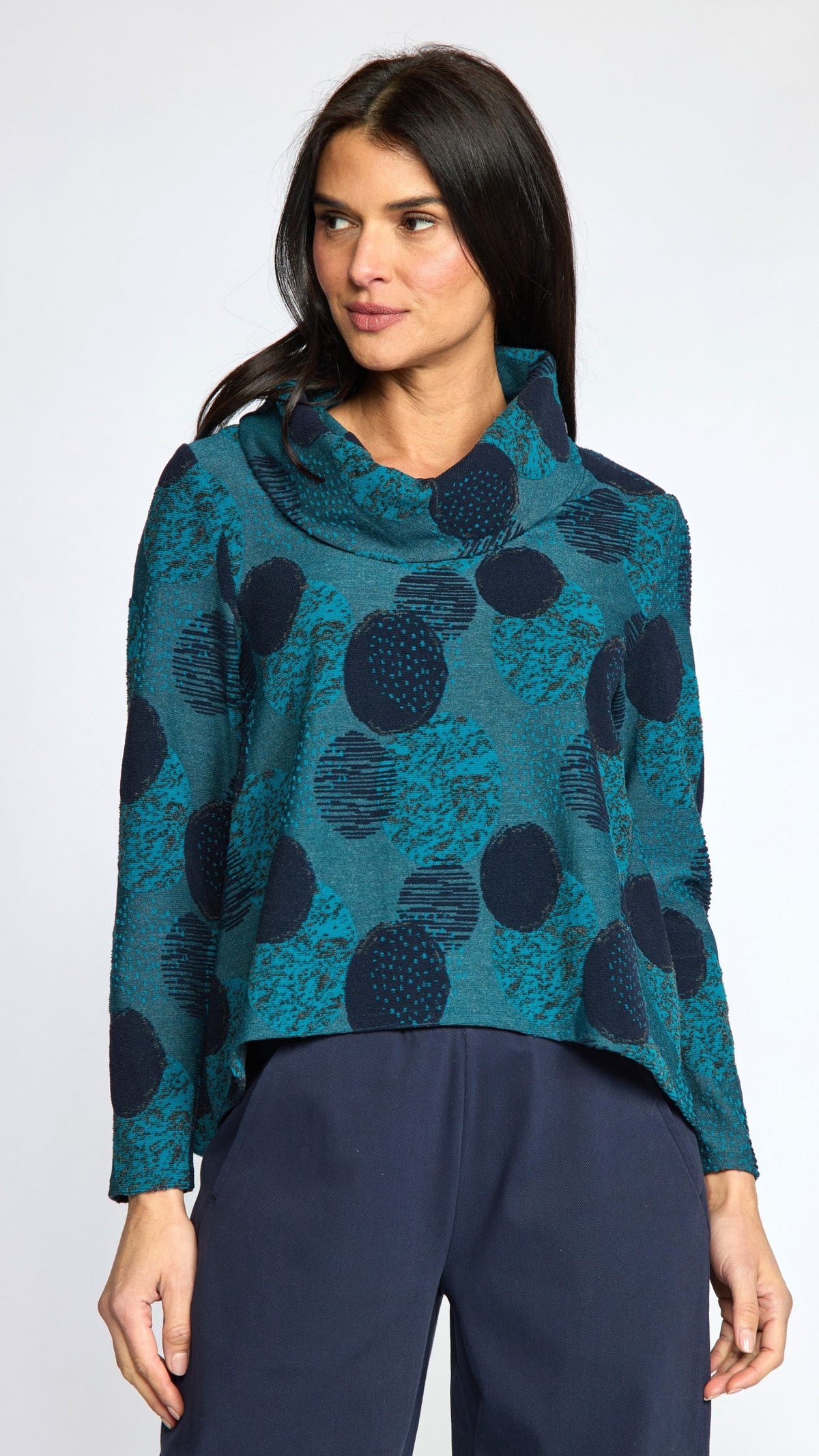 A woman modeling a teal and navy blue patterned cowl-neck long-sleeve top with dark navy pants, shot against a light gray studio background.