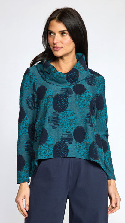 A woman modeling a teal and navy blue patterned cowl-neck long-sleeve top with dark navy pants, shot against a light gray studio background.
