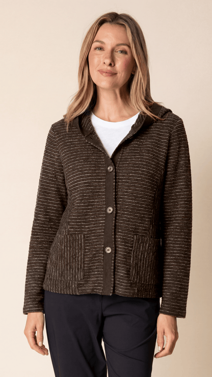 A woman modeling a brown textured knit cardigan with a hood and front buttons, worn over a white top and dark pants, against a solid beige background.