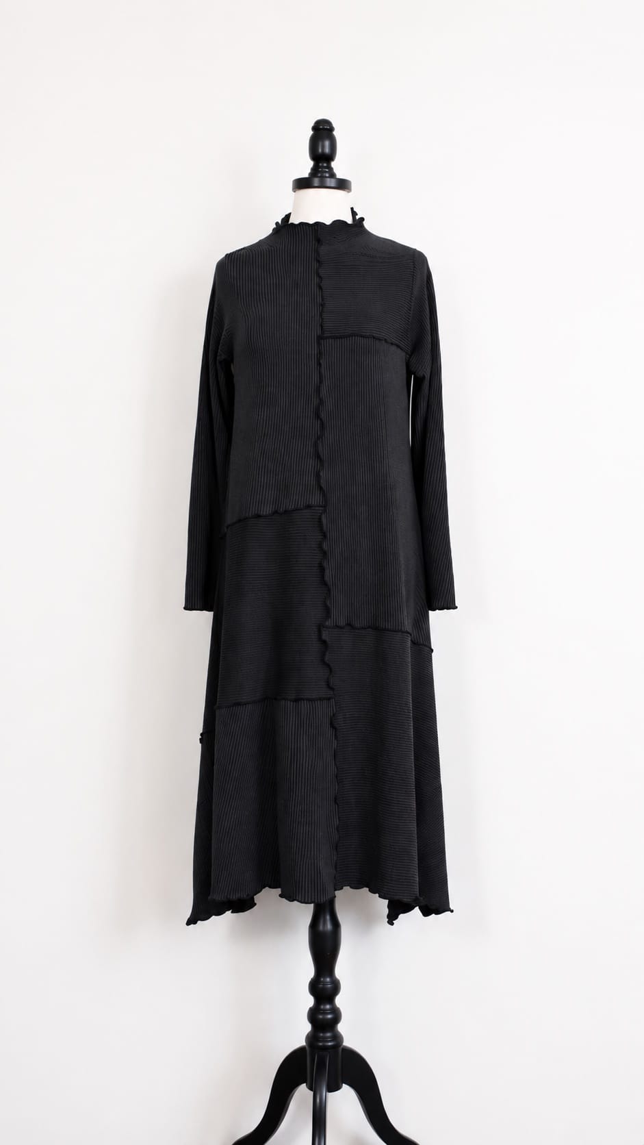 A black long-sleeve dress displayed on a black mannequin against a plain white wall. The dress features a high mock neck, a front asymmetrical zipper closure with ruffled trim, and a tiered skirt.