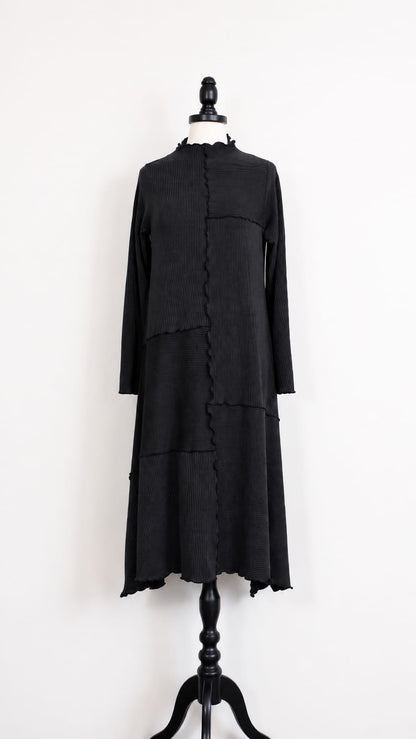 A black long-sleeve dress displayed on a black mannequin against a plain white wall. The dress features a high mock neck, a front asymmetrical zipper closure with ruffled trim, and a tiered skirt.