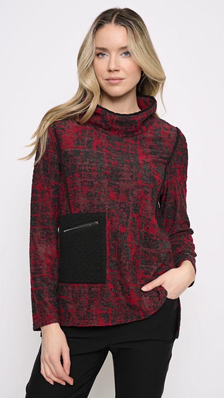 A woman modeling a red and black patterned mock neck sweater with a black front pocket, paired with black pants, against a plain white studio background.