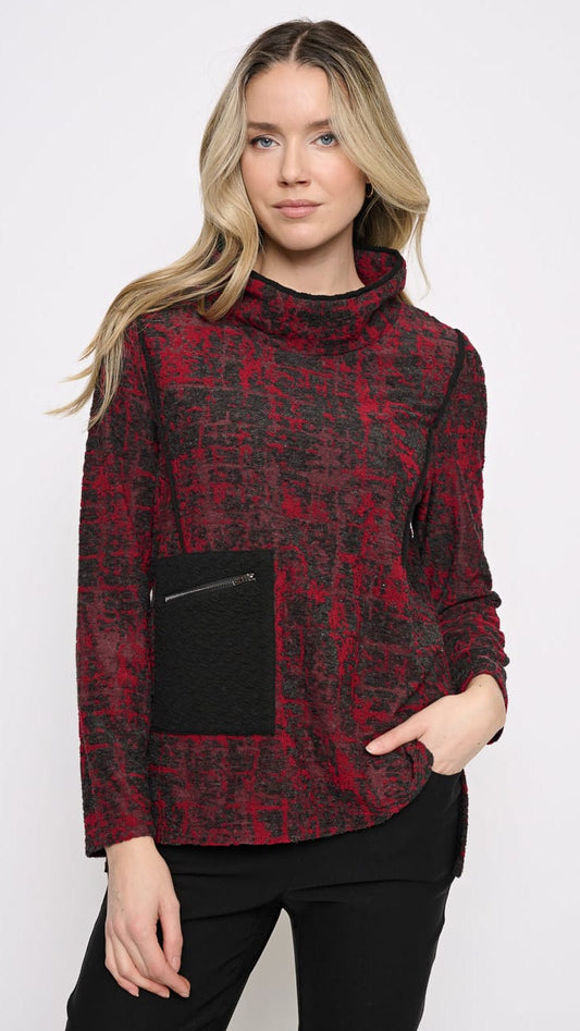 A woman modeling a red and black patterned mock neck sweater with a black front pocket, paired with black pants, against a plain white studio background.