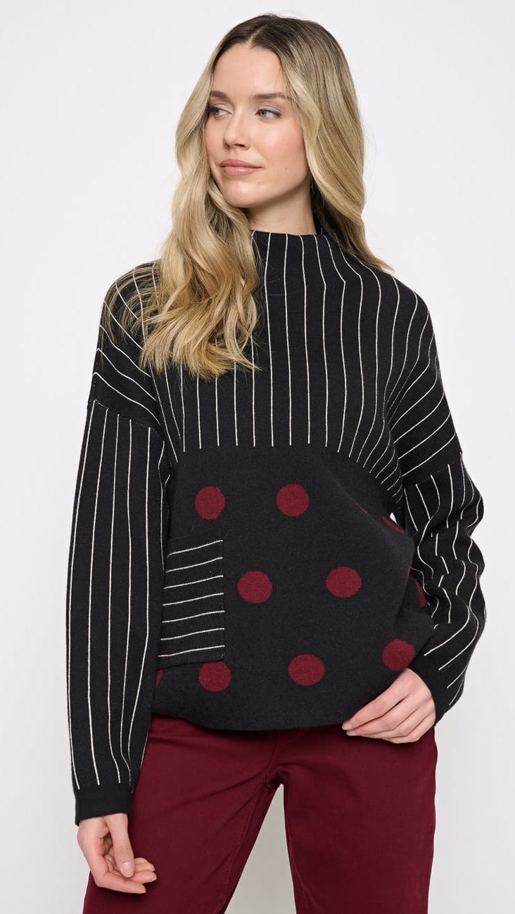 A woman modeling a black knit sweater with white pinstripes on the sleeves, a solid black front panel with red polka dots, and a striped pocket detail, paired with burgundy pants against a white studio background.