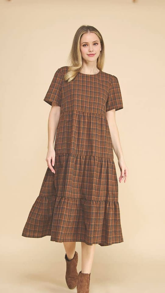 A woman modeling a short-sleeve tiered midi dress with a brown and orange plaid pattern, paired with brown ankle boots, against a solid beige studio background.