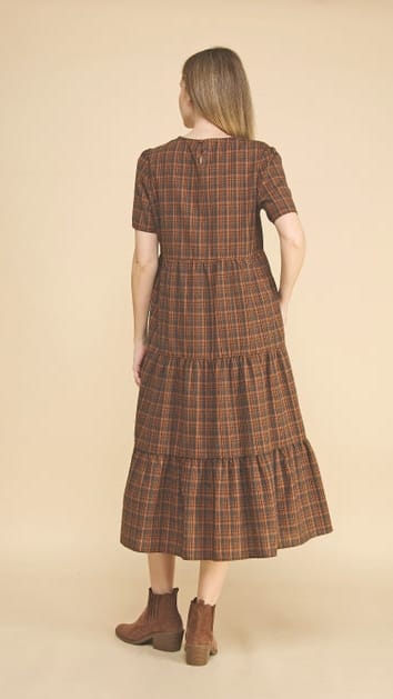 A woman shown from the back wearing a brown and tan plaid tiered midi dress with short sleeves, paired with brown ankle boots, against a solid beige studio background.