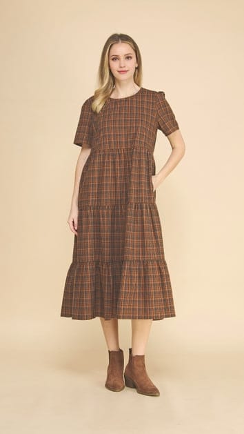 A woman modeling a brown and beige plaid midi dress with short sleeves, a round neckline, and a tiered skirt, paired with brown ankle boots, against a solid beige background.