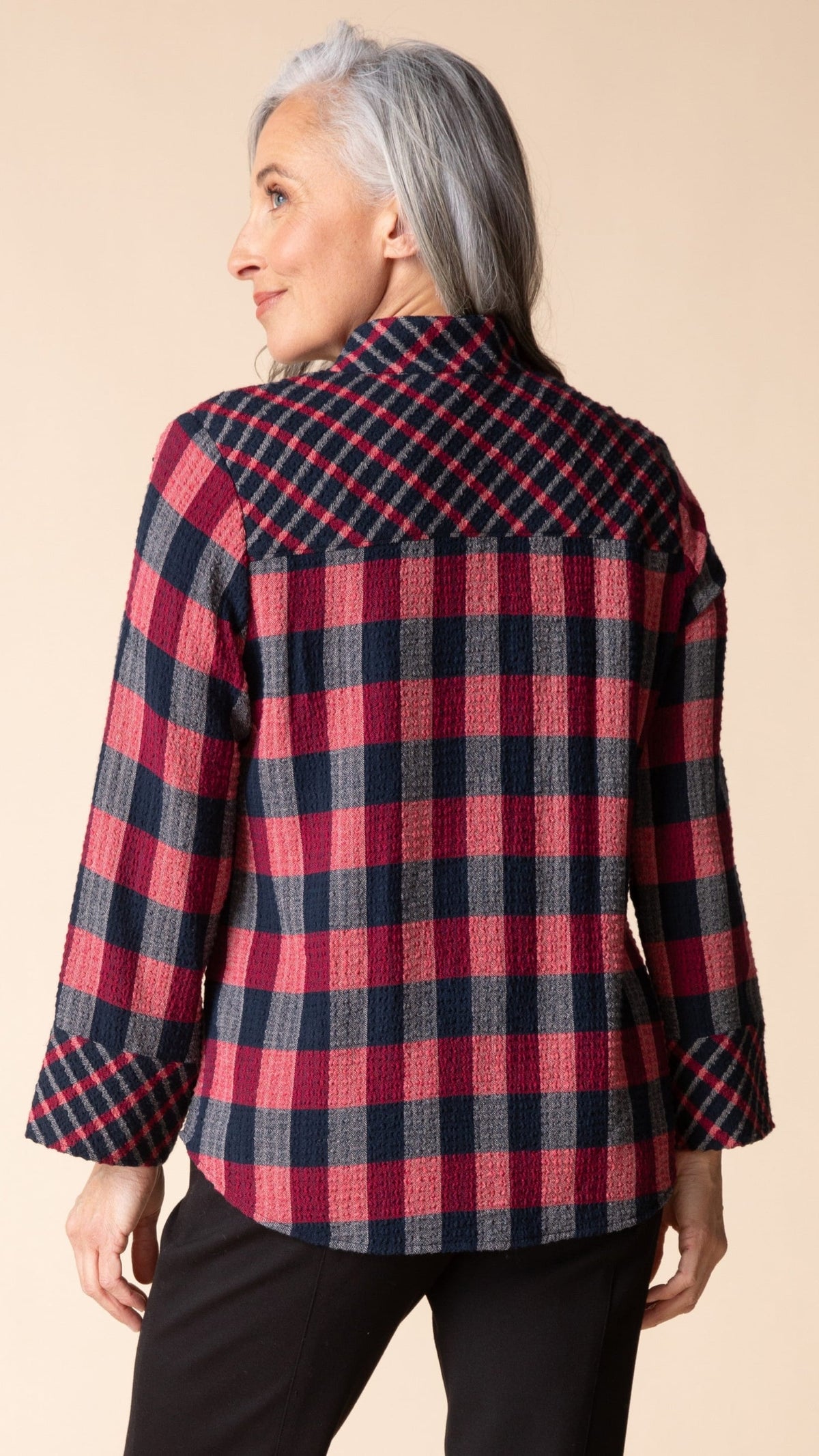 A woman with gray hair is shown from the back wearing a red, navy, and gray plaid flannel shirt. The photo is taken against a solid light beige background.